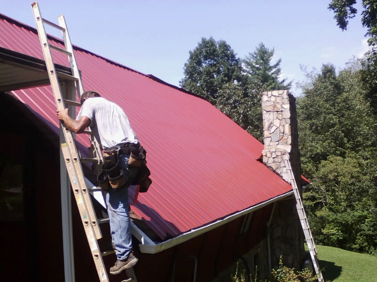 Roofer on a ladder beside a red metal roof and stone chimney — Soffit & Fascia Repair by Roofing Brothers in Pequea